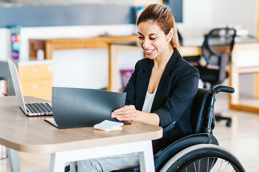 Client Center - Young Woman in a Wheelchair Works at a Desk, Checking a Folder of Papers and Smiling