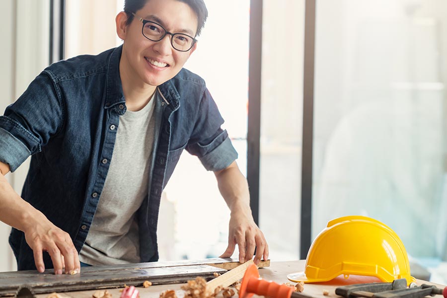 Specialized Business Insurance - Man in Denim Shirt and Glasses Smiles as He Measures Materials at a Drafting Table, Hard Hat Nearby